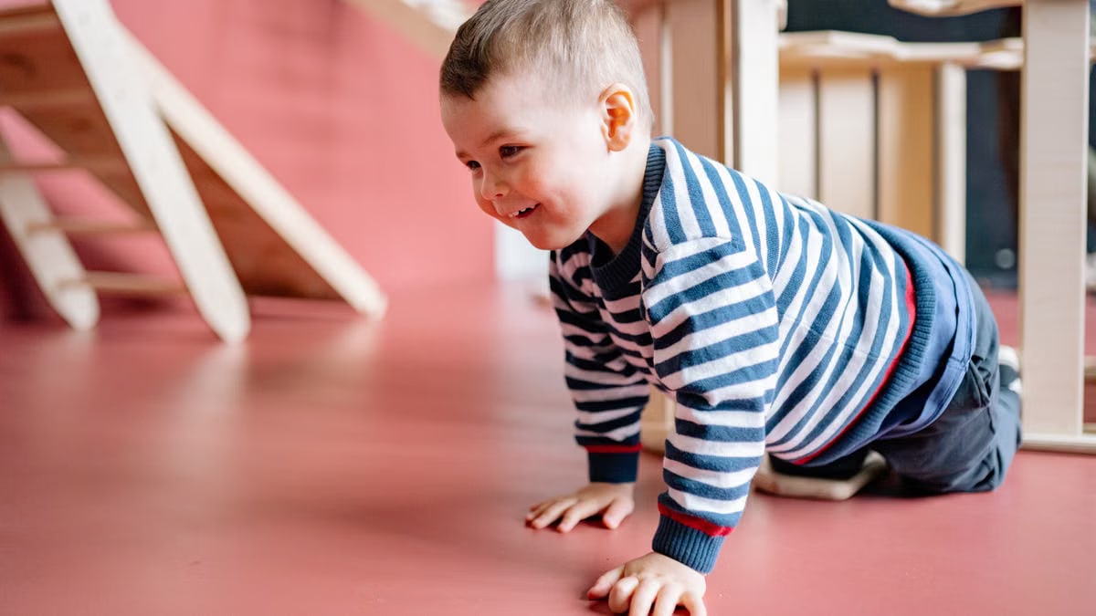 A cheerful toddler in striped long sleeves crawls on a red floor indoors, radiating joy.